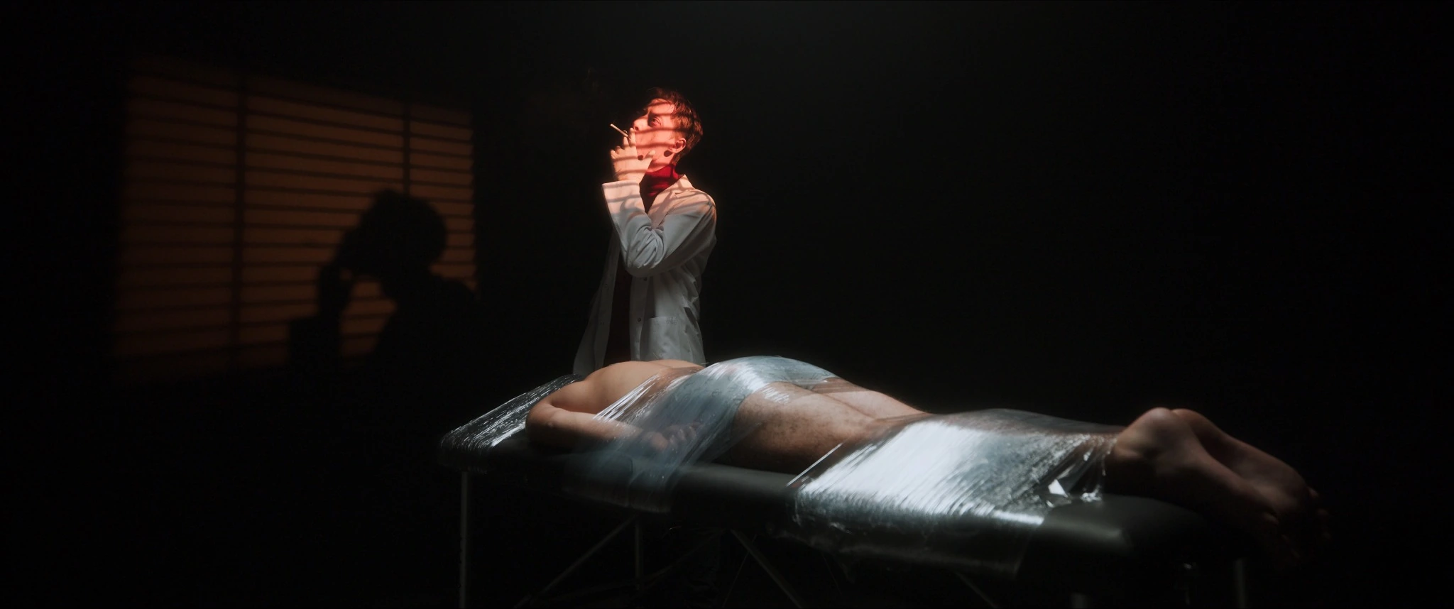 Warm slatted light on a man in a white coat beside a covered figure in Fluturon Fluturon